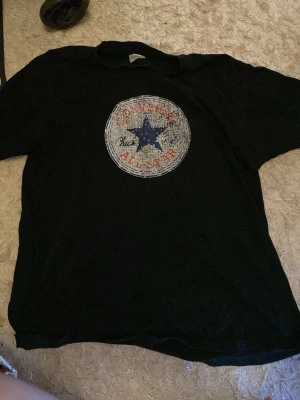 Svart Converse All Star t-shirt - Svart t-shirt från B&C Collection med stort Converse All Star-tryck i rött, blått och vitt på bröstet. Klassisk rund hals och korta ärmar. Perfekt för dig som gillar streetwear och vill ha en ikonisk look.