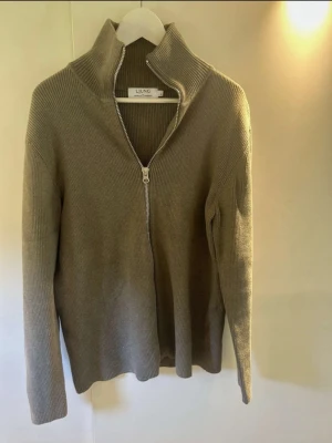 Beige ribbad kofta från Ljung - Säljer en beige ribbad kofta från Ljung med hög krage och dragkedja framtill. Tröjan är långärmad och har en clean, stilren look som passar perfekt till lager på lager under höst och vinter.