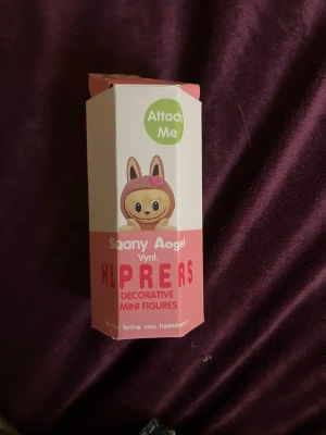 Sonny angel med labubu ansikte  - Bilderna visar en dekorativ mini figur från Sonny angel, med labubu ansikte. Figurerna har olika färger på huvudet och djuröron, men ingen väst eller liknande plagg är synlig. 