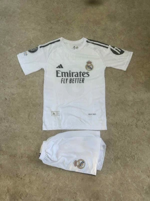 Real Madrid Mbappé matchställ - Säljer ett Real Madrid matchställ med Mbappé och nummer 9 på ryggen. Setet består av vit tröja med svarta detaljer och shorts med klubbens emblem. Materialet är lätt polyester och har HEAT.RDY-teknologi för extra komfort. Perfekt för fotbollsträning eller att bära på läktaren.