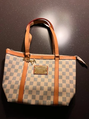 Louis Vuitton handväska - Säljer en snygg handväska från Louis Vuitton med klassiskt rutigt mönster i beige och grått. Väskan har ljusbruna skinnhandtag och detaljer, samt en metallplatta med logga framtill. Perfekt storlek för att få plats med allt du behöver och stängs med dragkedja.