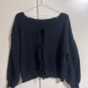 Svart stickad kofta med snörning - Svart stickad kofta med långärmade, lite puffiga ärmar och snygg snörning framtill. Koftan har en rundad hals och ribbade muddar vid ärmslut och nederkant. 