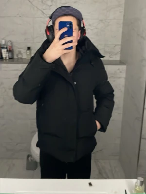 Svart pufferjacka från Canada Goose - Säljer en svart pufferjacka från Canada Goose med huva och diskret logga på ärmen. Jackan är vadderad och har en clean look med dragkedja och knappar framtill. Perfekt för kalla dagar och riktigt skön att bära. Snygg och enkel att matcha med allt.