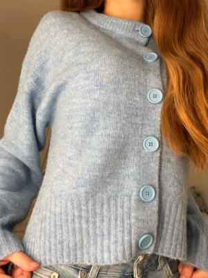 Ljusblå stickad kofta med knappar - Mysig ljusblå stickad kofta med stora matchande knappar framtill och ribbad nederkant. Koftan har lång ärm och en cool slits på sidan som ger extra stilpoäng. Perfekt att slänga på över en t-shirt och jeans för en chill look.