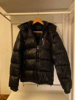 Svart pufferjacka från Polo Ralph Lauren - Svart pufferjacka från Polo Ralph Lauren med huva och dragkedja framtill. Jackan har ett broderat rött Polo-logo på bröstet. Fylld med dun och har en glansig finish, perfekt för kalla dagar.