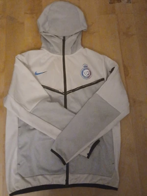 Nike tech Al nassr kofta - Säljer en stilren vindjacka från Nike i ljusgrått och vitt med svart dragkedja och huva. Jackan har Al Nassr FC-logga på bröstet och blå Nike-swoosh. Tillverkad i 100% polyester, perfekt för sport och chill. Långärmad och med praktiska fickor. Köpte för 799kr men säljer nu för 499kr
