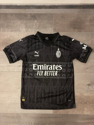 Svart AC Milan x Puma t-shirt - AC Milan x Puma t-shirt i svart med grått grafiskt mönster inspirerat av gotisk arkitektur. Kortärmad med krage och tryckta loggor från Puma, Pleasures och Emirates på framsidan. Materialet är lätt polyester, perfekt för fotboll eller häng med kompisar.