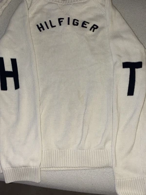 Vit stickad tröja Tommy Hilfiger - Säljer en vit stickad tröja från Tommy Hilfiger i storlek L barn storlek. Tröjan har svarta bokstäver på bröstet och ärmarna, samt ribbade muddar vid ärmslut och nederkant. Perfekt för dig som gillar klassisk och stilren design med tydlig logga.