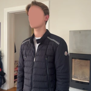 Mörkblå Moncler pufferjacka herr - Mörkblå pufferjacka från Moncler med quiltad design, ståkrage med knapp, tvåvägsdragkedja och vita detaljer på axlarna. Jackan har Moncler-logga på vänster ärm och två dragkedjefickor framtill. Perfekt för dig som vill ha en clean och stilren look. Köpt från Care of Carl och kvitto finns! Pris:3999 13-16 år!