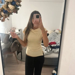 Ljusgul figurnära topp med drapering - Säljer en ljusgul topp med korta ärmar och snygg drapering vid halsen. Toppen är figurnära och har en mjuk, stretchig känsla som sitter snyggt på kroppen. Perfekt att styla med jeans eller kjol för en fräsch look.