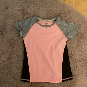 Sportig rosa tränings-tröja från H&M Sport - Sportig tränings-tröja från H&M Sport i ljusrosa med gråmelerade korta ärmar och svarta paneler på sidorna. Perfekt för träning eller aktiva dagar. Tillverkad i ett stretchigt syntetmaterial som andas och ger bra rörelsefrihet.