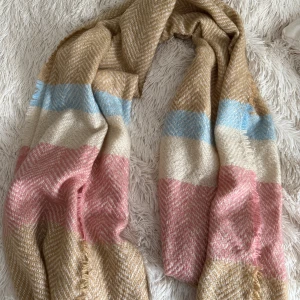 Randig halsduk från Accessories - Supermjuk och fluffig halsduk med breda ränder i beige, ljusblått och rosa. Stickad struktur med fransar i kanterna, perfekt att svepa runt halsen för extra värme. Snygg och färgglad accessoar som lyfter vilken outfit som helst.