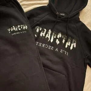 Trapstar dress - Säljer en svart hoodie från Trapstar med fet broderad logga och texten 'IT'S A SECRET' på bröstet. Hoodien har snörning i huvan och är gjord i mjuk bomullsmix. Perfekt för dig som gillar streetwear och vill sticka ut med en ikonisk look.