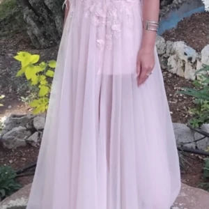 Ljusrosa långklänning med spetsdetaljer - Superfin ljusrosa långklänning med tunna axelband och transparent, flowy kjol. Överdelen har vackra spets- och broderidetaljer samt 3D-blommor. Klänningen har slits framtill och öppen rygg, vilket ger en romantisk och elegant vibe. Perfekt för speciella tillfällen! Använd en kväll. Tvättad. 