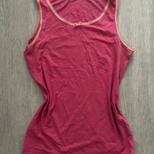 Rosa linnetopp med spetskant och rosett - Söt rosa linnetopp med ljusrosa spetskant runt halsringning och ärmhål. Dekorerad med en liten rosett framtill. Toppens material är mjuk bomull och modellen är ärmlös med rundad hals. Perfekt för en avslappnad och feminin look. xs/s