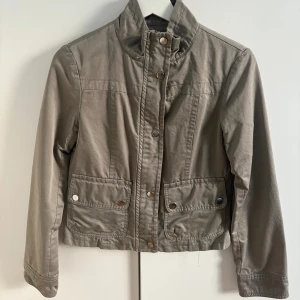 Grön  vindjacka med fickor - Snygg grön vindjacka med hög krage och tryckknappar framtill. Jackan har två stora fickor, samt långa ärmar. 