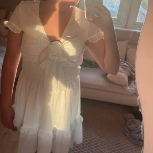 Vit kort omlottklänning med volanger - Supersöt vit kort klänning med omlottknytning framtill och bred smock i midjan. Klänningen har volangdetaljer nertill och på ärmarna, samt korta ärmar med lätt rynk. Perfekt för sommarens alla tillfällen! Medium aldrig använd 