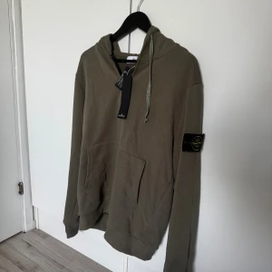 Olivgrön hoodie från Stone Island - Snygg olivgrön hoodie från Stone Island med klassisk patch på ärmen. Tröjan har huva med snörning, känguruficka och långa ärmar. Perfekt för dig som gillar streetwear och vill ha en stilren look.