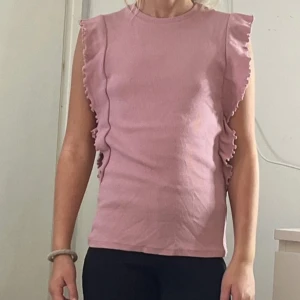 Rosa linnetopp med volangdetaljer - Säljer en ljusrosa linnetopp med volangdetaljer längs axlarna och sidorna. Toppen är ärmlös och har en rund halsringning. Materialet är mjukt och känns som bomull, perfekt för sommarens alla dagar. Enkel och söt design som ger en feminin vibe.