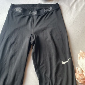 Svarta Nike  träningsbyxor  - Svarta leggings från Nike med Dri-Fit-teknologi och elastisk midja med Nike-logga runtom. Swoosh-logga på benet. Perfekta för träning och tillverkade i ett stretchigt syntetmaterial som andas.