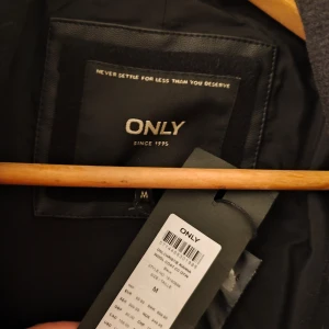 Svart ullkappa från ONLY med bälte - Stilren svart ullkappa från ONLY i storlek M. Kappan har hög krage, dragkedja och knappar framtill, samt praktiska fickor med dragkedja. Bälte i midjan för snygg passform och långa ärmar med dragkedjedetaljer vid ärmslut. Perfekt för kyliga dagar.