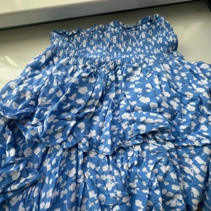 Blommig blå volangkjol Samsa Collection - Supersöt blå kortkjol från Samsa Collection med vita blommor och volanger. Kjolen har smock i midjan för skön passform och är gjord i mjuk bomull. Perfekt för dig som gillar färgglada och lekfulla plagg med mycket rörelse. Längd = 42 cm har en liten fläck på framsidan 