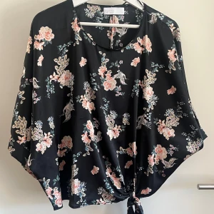 Svart blommig blus från Samsa Collection - Svart blus med blommigt mönster i rosa och beige toner från Samsa Collection. Blusen har vid passform, korta ärmar och en snygg knytning framtill. Materialet är mjukt och har en lätt glansig yta som ger en lyxig känsla. 