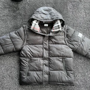 Burberry vinterjacka - Svart pufferjacka från Burberry med avtagbara ärmar så du kan bära den som väst. Jackan har huva, dragkedjor på fickorna och ett rutigt foder i grått, vitt och rött. Märkespatch på ärmen och klassisk Burberry-label på insidan. Perfekt för kalla dagar. Skick 10/10 som ny, storlek L, pris går att diskutera✅