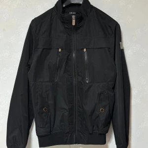 Svart bomberjacka från SMOG (HELT OANVÄND) - Svart bomberjacka från SMOG med klassisk krage, dragkedja framtill och flera fickor med dragkedjor och detaljer i metall. Jackan har muddar vid ärmslut och midja. Perfekt för dig som gillar en clean och stilren look. Storlek S. 