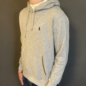 Grå hoodie från Ralph Lauren - Säljer en stilren grå hoodie från Ralph Lauren med klassisk broderad logga på bröstet. Tröjan har huva med snörning, magficka och ribbade muddar. Perfekt för chill dagar och enkel att matcha med jeans eller joggers.