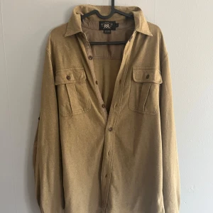 Beige overshirt från Ralph Lauren RRL - Snygg grön/beige overshirt från Ralph Laurens exklusiva dottermärke RRL. Skjortan har klassisk krage, två bröstfickor med lock och knappar framtill. Perfekt för lager-på-lager och chill stil. Passar till alla möjliga stilar.