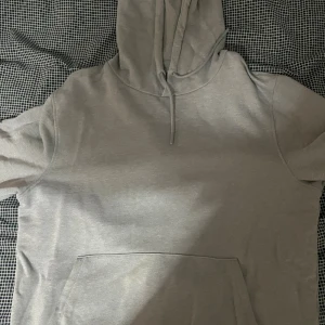 Grå hoodie från Dressmann i bomull - En stilren grå hoodie från Dressmann i mjuk bomull. Tröjan har huva med snörning och en stor magficka framtill. Perfekt för chill dagar eller när du vill ha en enkel och clean look. Klassisk passform och långärmad modell. Tröjan är i storlek M. 