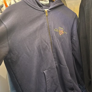 blå hoodie från Gant med zipper - Snygg marinblå hoodie från Gant med broderad logga på bröstet och hel dragkedja framtill. Tröjan har huva, känguruficka och är tillverkad i mjuk bomull. Perfekt för en chill och sportig stil. Använd men fortfarande i fantastiskt skick.