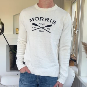 Vit stickad tröja från Morris Club - Vit stickad tröja från Morris Club med svart tryck framtill och ribbade muddar vid ärmslut och nederkant. Klassisk rund halsringning och snygg passform, perfekt till jeans. Stort Morris-tryck och korsade åror på bröstet.