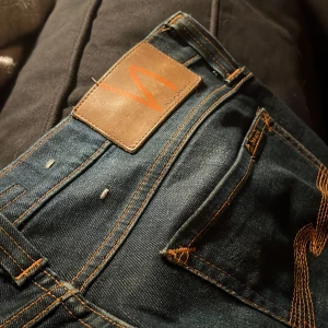  Nudie Jeans - Säljer ett par mörkblå jeans från Nudie Jeans med klassisk femficksdesign och snygga orangea sömmar. Jeansen har en rak passform och är tillverkade i slitstark bomullsdenim. Märkespatch i läder baktill och coola detaljer på bakfickorna.