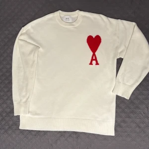 Vit stickad tröja från Ami Paris - Vit stickad tröja från Ami Paris med röd hjärtlogga och bokstaven A på bröstet. Klassisk rund halsringning och ribbade muddar vid ärmslut och nederkant. Perfekt för dig som gillar stilrena plagg med en twist. Helt oanvänd.