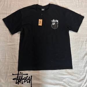 Svart Stüssy t-shirt med 8-ball print - Svart t-shirt från Stüssy i 100% bomull med klassisk logga och stor 8-ball print på bröstet. Skön passform och streetwear-vibe, perfekt för dig som gillar snygga och enkla statement-plagg.