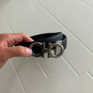 Svart skärp från Ferragamo i skinn - Säljer ett svart skärp från Ferragamo i äkta skinn med en ikonisk, stor silverspänne i metall. Bältet har stilrena sömmar längs kanterna och klassisk design som ger en lyxig känsla. Perfekt accessoar för att lyfta din outfit.