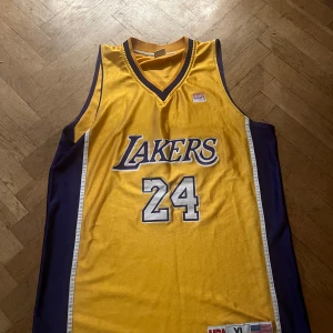 Lakers basketlinne Kobe Bryant #24 - Gult och lila basketlinne från Lakers med nummer 24 och Bryant på ryggen. Klassisk v-ringad modell med breda axelband och tryckta detaljer. .