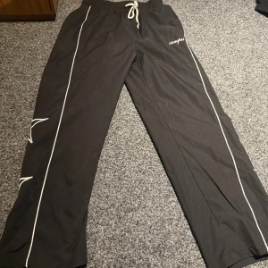 Svarta campusbyxor med stjärnor - Svarta byxor från Campus med vita stjärnor broderade längs benen och vita ränder på sidorna. Byxorna har snörning i midjan och är tillverkade i ett mjukt syntetmaterial. Perfekt för dig som gillar en chill och unik stil.