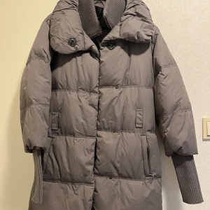 Grå dunjacka från Amisu, strl 38 - Säljer en grå dunjacka från Amisu i storlek 38. Jackan har quiltad design, stora knappar framtill, ribbade muddar och hög krage. Perfekt för kalla dagar med sin långa modell och praktiska fickor.