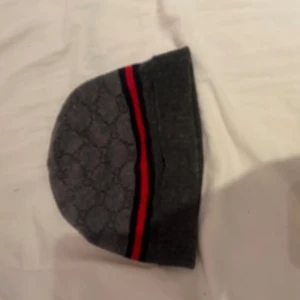 Gucci grå mössa i ull och kashmir - Gucci mössa i grått med diskret GG-mönster och en röd samt svart rand runt kanten. Tillverkad i 70% ull och 30% kashmir för en lyxig och mjuk känsla. Perfekt accessoar för att lyfta din vinterstil.
