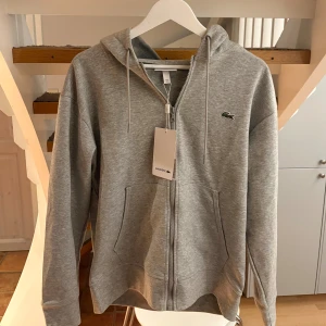 Grå hoodie från Lacoste med dragkedja - Snygg grå hoodie från Lacoste i klassisk passform. Tröjan har huva med snörning, dragkedja framtill och två fickor. Ikoniska Lacoste-loggan på bröstet. Perfekt för chill dagar eller när du vill ha en clean streetstyle.