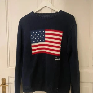 Marinblå stickad tröja från Gant - Snygg marinblå stickad tröja från Gant med amerikansk flagga i rött, vitt och blått på bröstet. Rund hals och långa ärmar, perfekt för dig som gillar klassisk preppy stil. Gant-logga broderad under flaggan.
