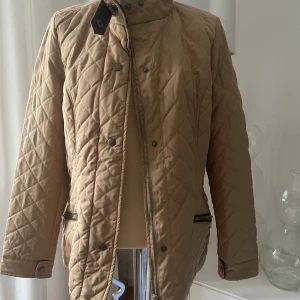 Beige quiltad jacka från Abacus  - Säljer en beige quiltad jacka från Abacus med snygga detaljer som dragkedja, knappar och två fickor med dragkedja. Jackan har hög krage och lång ärm, perfekt för dig som gillar klassisk men trendig stil. Oanvänd 
