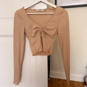 Beige croppad topp från NA-KD - Säljer en beige croppad topp från NA-KD med långa ärmar och v-ringning. Toppen har ribbad struktur och snygg snörning framtill som ger en cool detalj. Perfekt att matcha med höga jeans eller kjol för en trendig look.