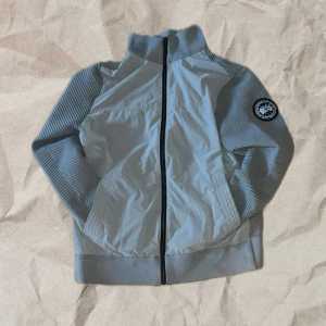 Canada Goose cardigan  - Snygg grå cardigan från Canada Goose med ribbade ärmar och hög krage. Jackan har hel dragkedja framtill, två fickor och en svartvit logotyp på ena ärmen. Materialmix med stickade partier och slät framsida ger en modern look. Den passar på folk mellan 165-180cm. Inget tilbehör förekommer därav priset.