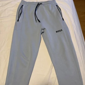 Ljusblå mjukisbyxor från BOSS - Snygga ljusblå mjukisbyxor från BOSS med svart logga på benet. Byxorna har elastisk midja med svart snörning och praktiska fickor med dragkedja. Perfekta för chill eller träning, mjukt material och relaxed passform.