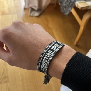 Christian Dior svart tygarmband - Säljer ett svart och vitt armband från Christian Dior med broderad logga och snygga tofsar i ändarna. Armbandet är gjort i tyg och har ett justerbart knytband för perfekt passform. Perfekt accessoar för att lyfta din outfit.