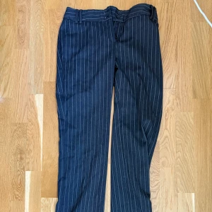 Randiga kostymbyxor från MNG Suit - Snygga mörkblå kostymbyxor från MNG Suit i storlek 36. Byxorna har ett klassiskt rakt snitt och är dekorerade med tunna vita ränder. De har hällor för bälte och stängs med knapp och dragkedja. Perfekta för en stilren look.